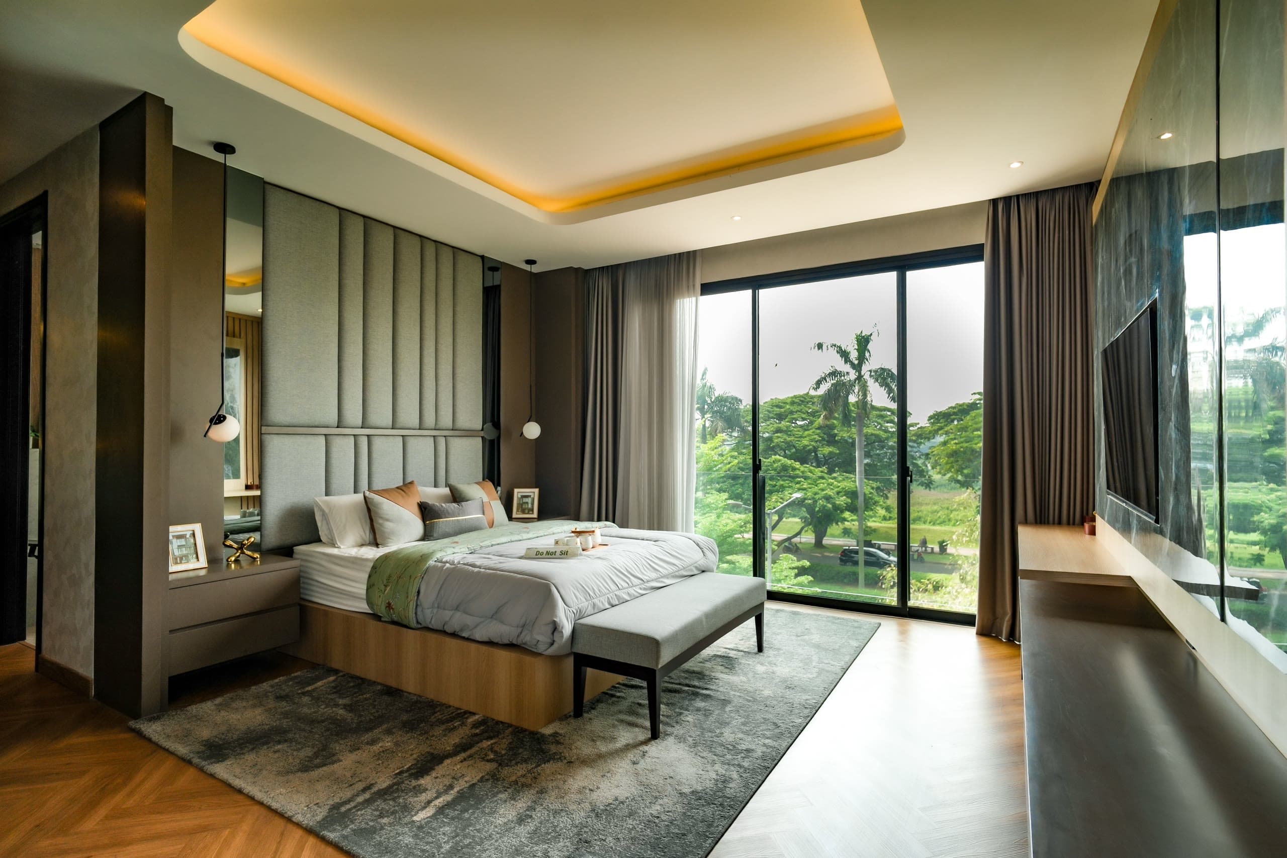 O mesmo quarto com cama de casal, cabeceira estofada, banco aos pés da cama, tapete e cortinas